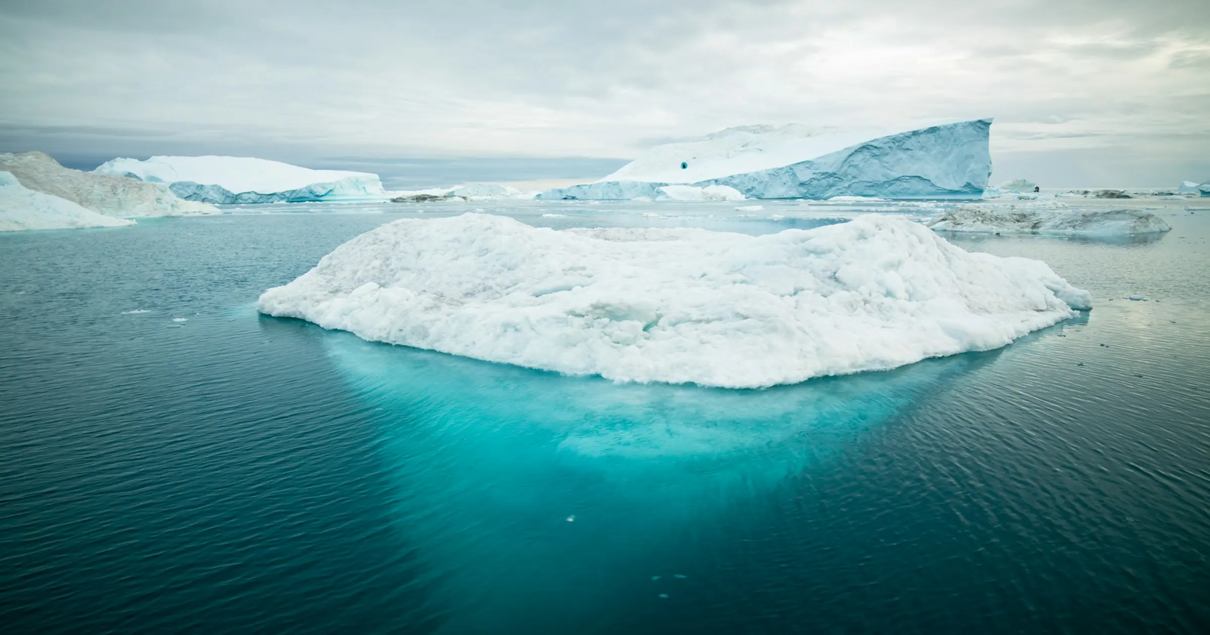 An iceberg in calm water with most of its bulk showing as a turquoise mass below the surface