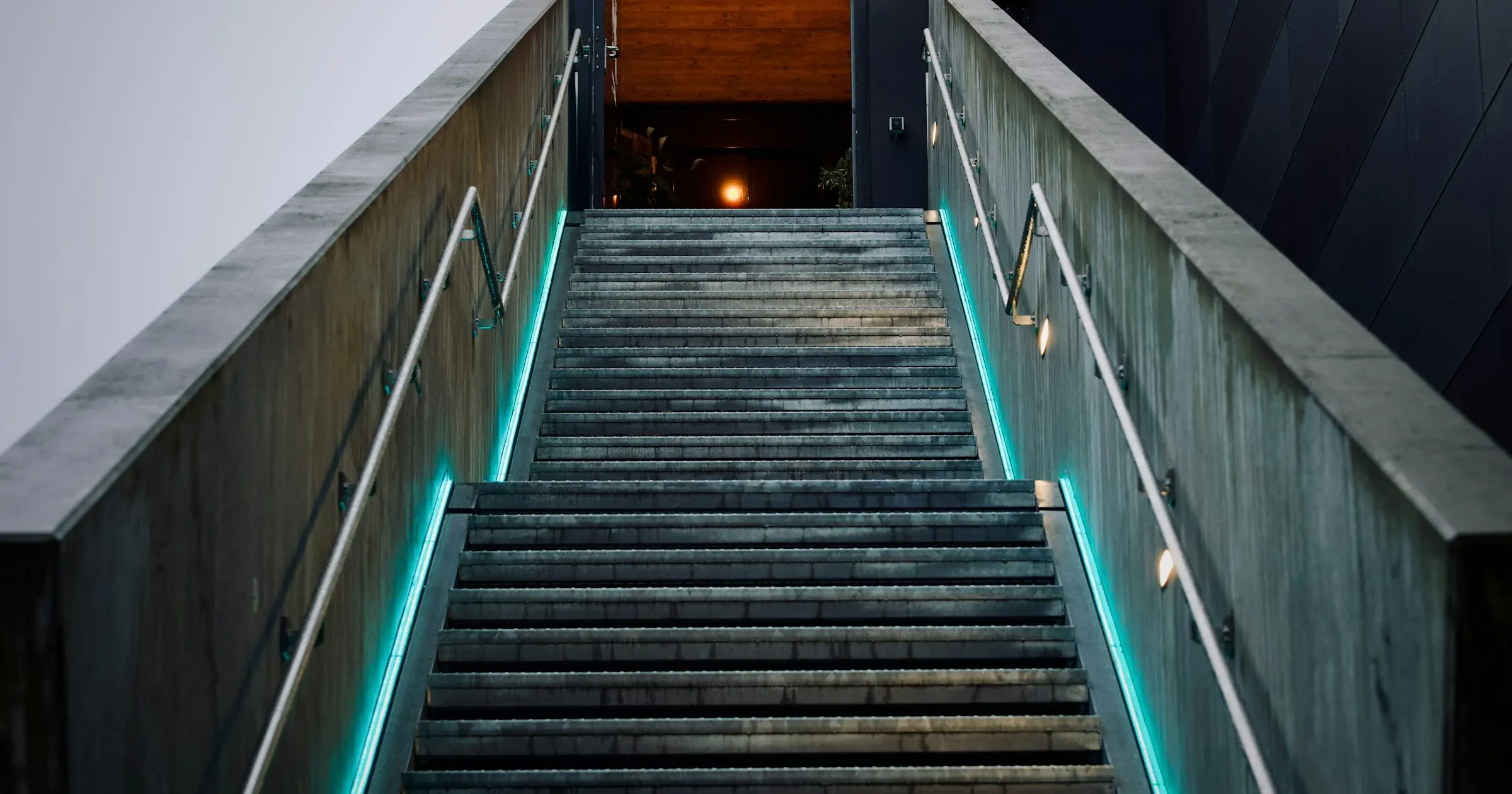 Concrete staircase with teal edge lighting climbing up toward a lit doorway