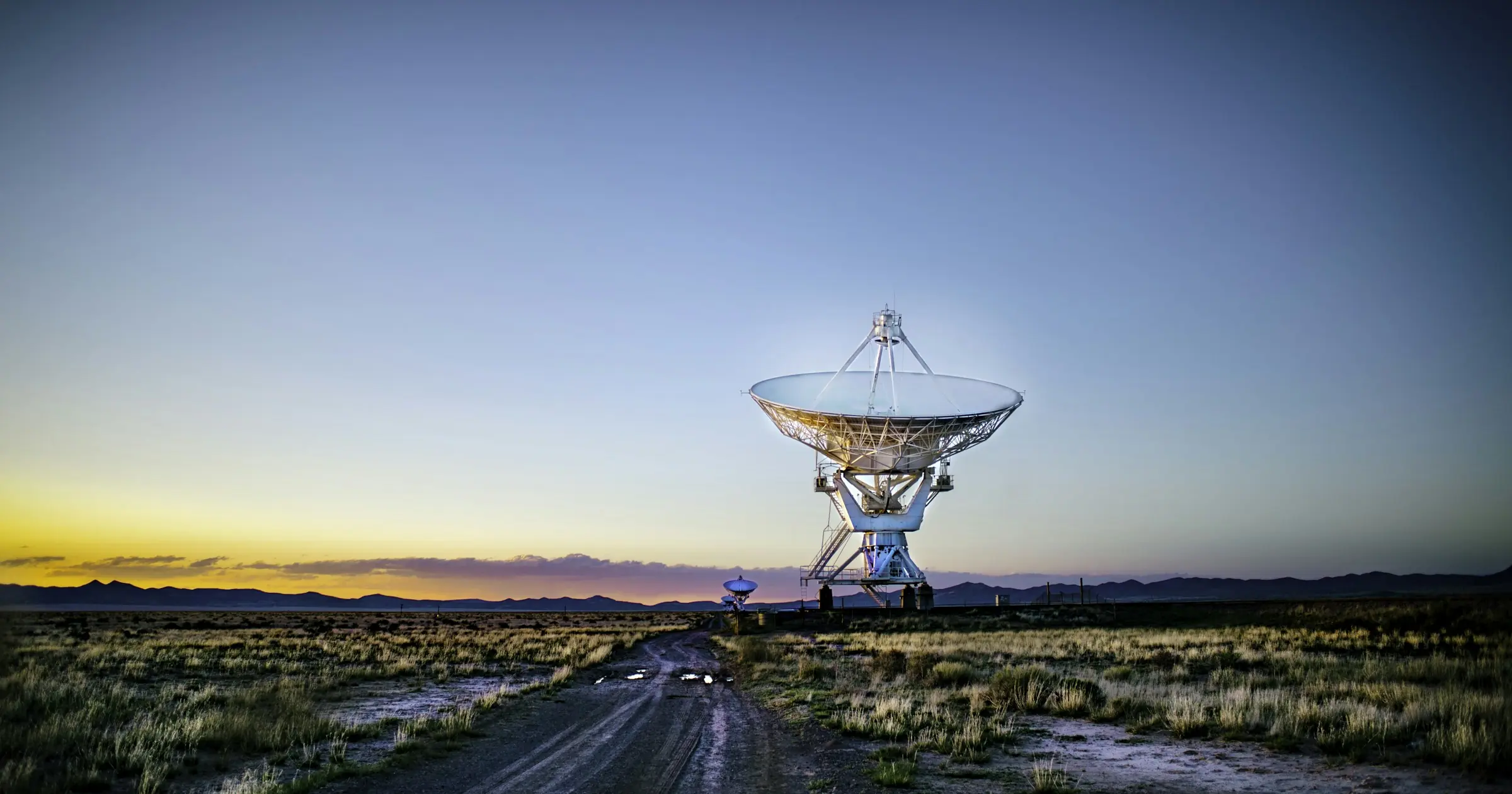 A large radio telescope dish at the end of a dirt road at dusk