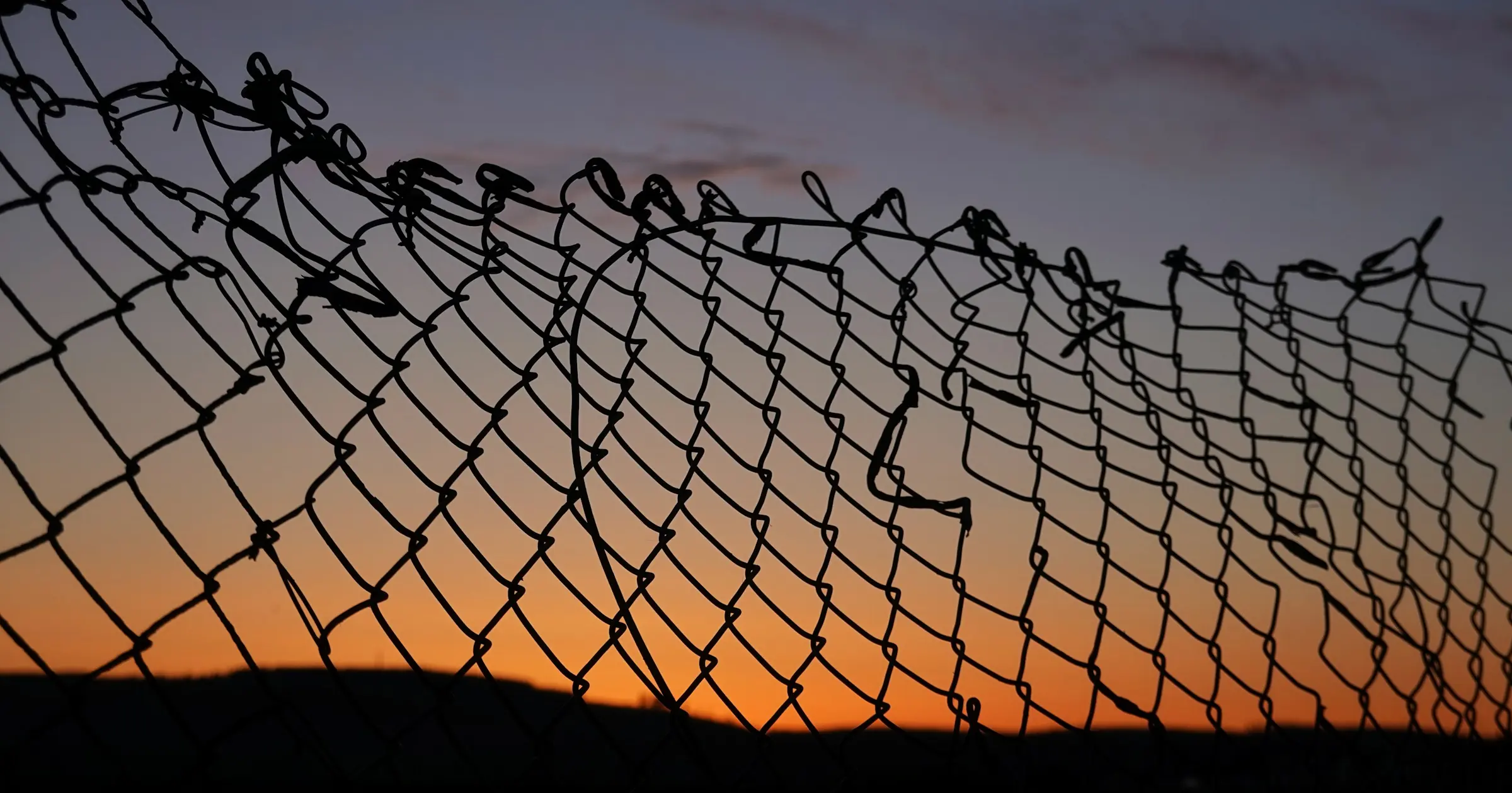A torn chain-link fence silhouetted against an orange sunset sky