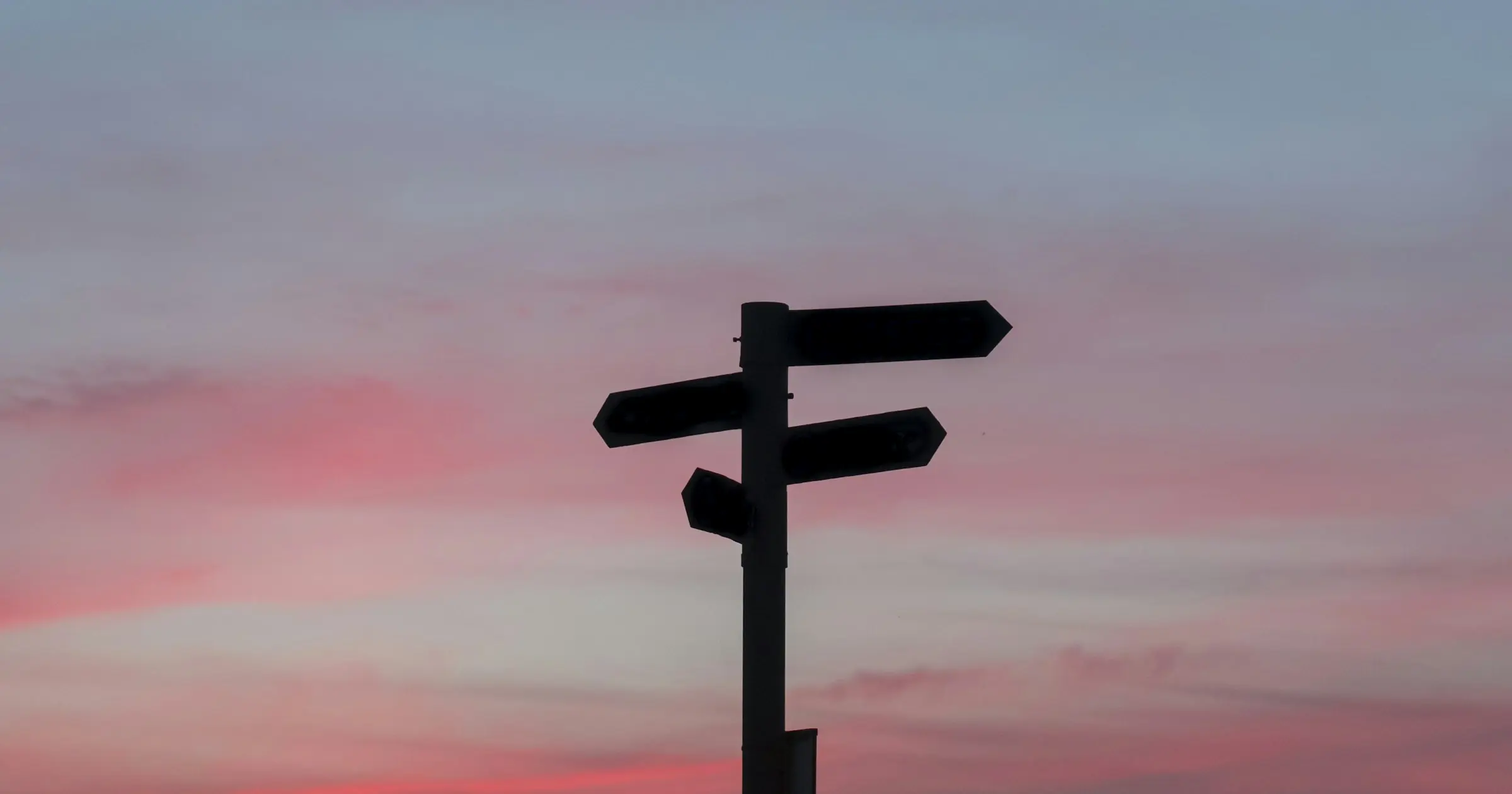 The silhouette of a signpost with blank arms pointing different ways against a pink sunset sky