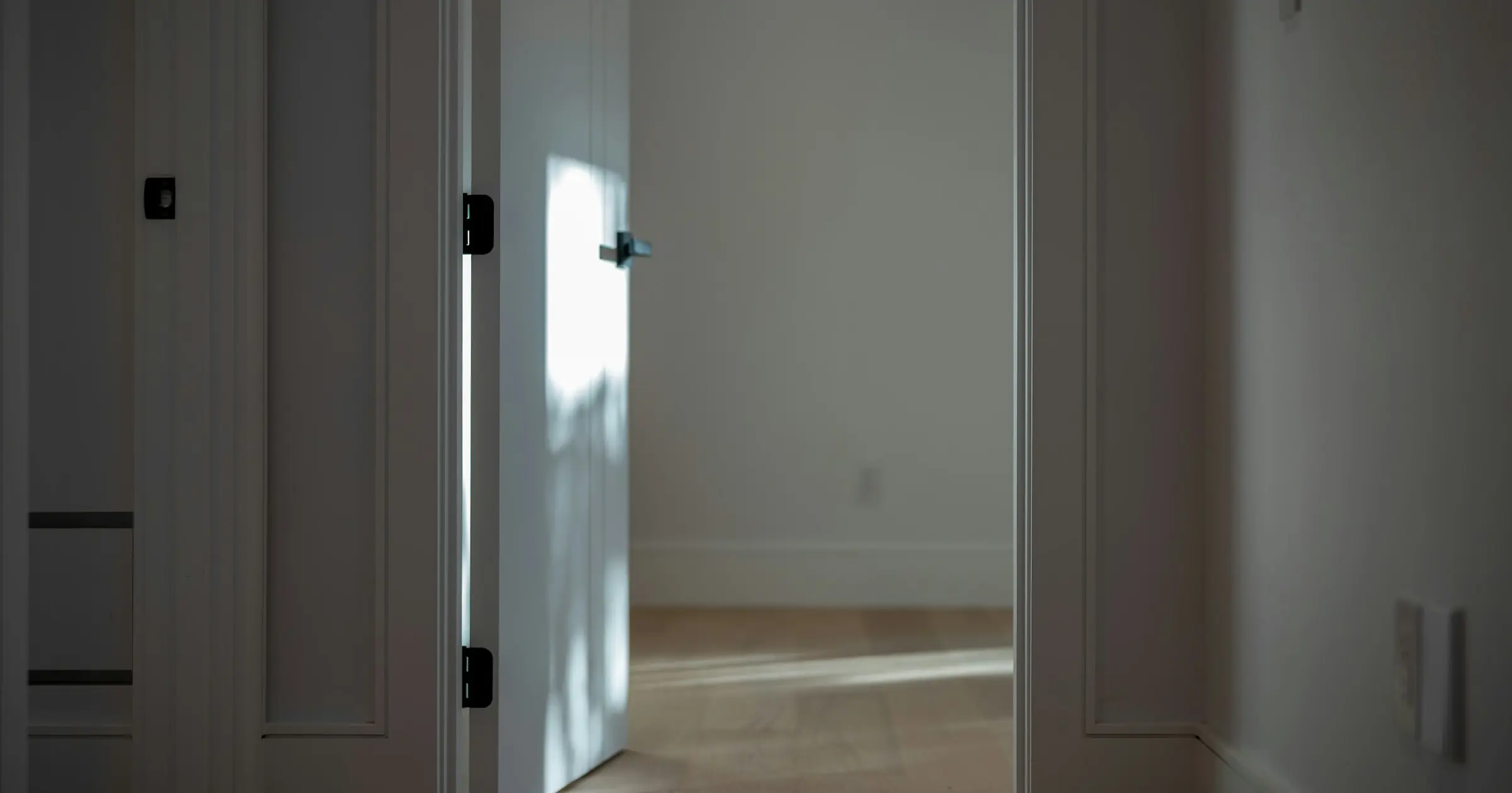 A white door standing half open onto an empty room with sunlight on the wooden floor