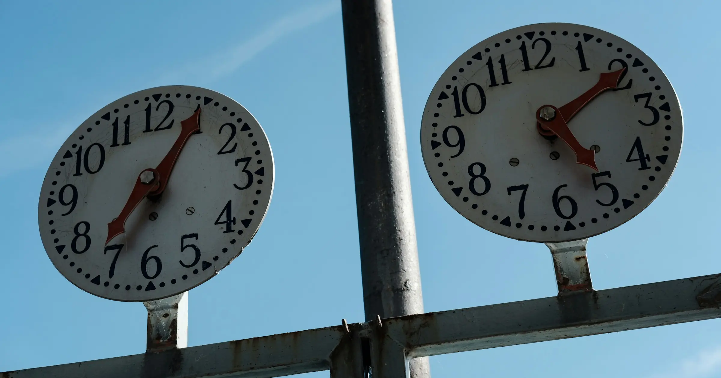 Two outdoor clocks mounted side by side on a metal pole, showing different times against a blue sky.