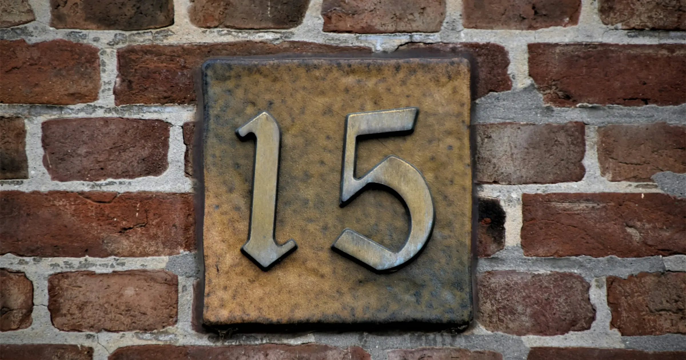 A bronze house number plate showing the number 15 on a brick wall.