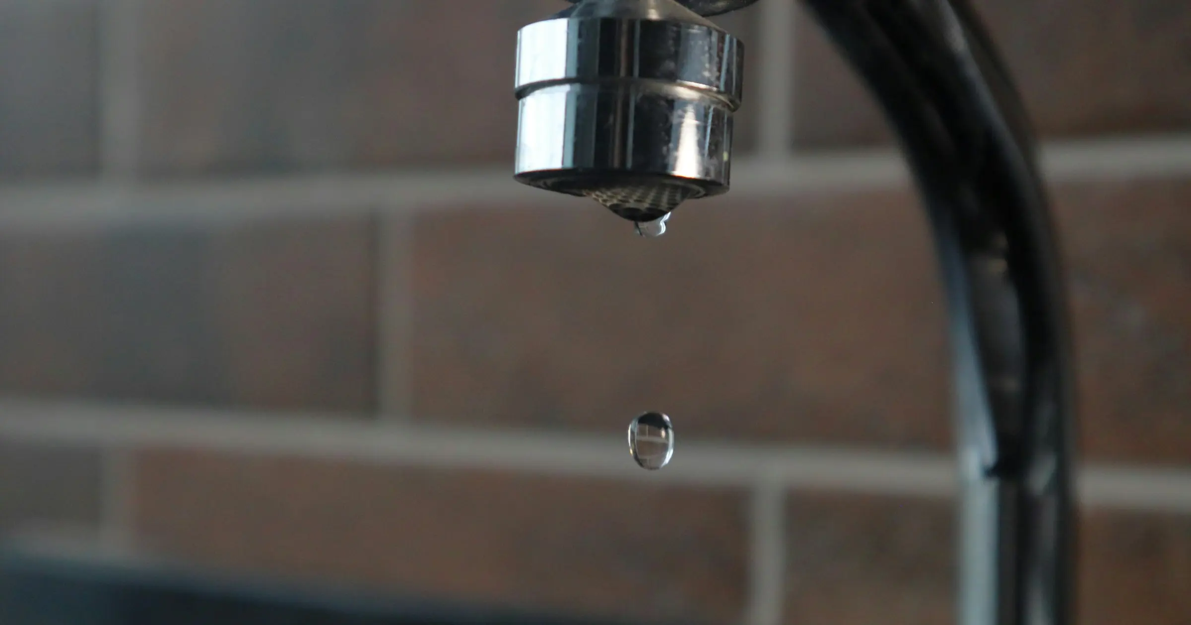 A single drop of water falling from a chrome tap in front of a brick wall