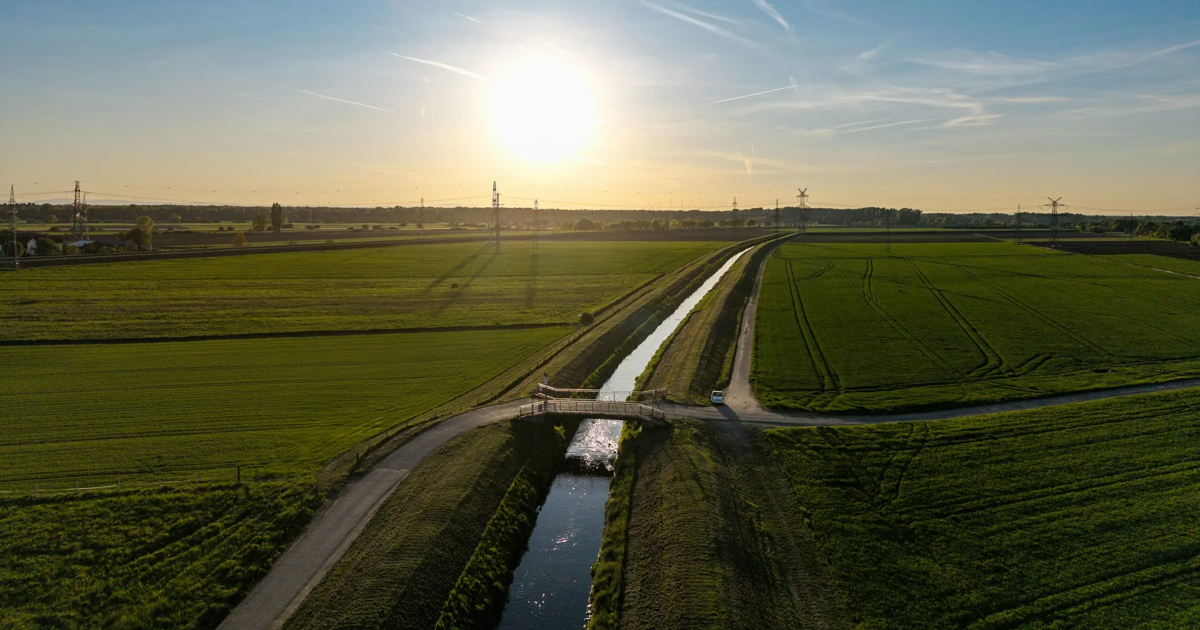 Aerial view of a straight canal running through green fields toward a low sun