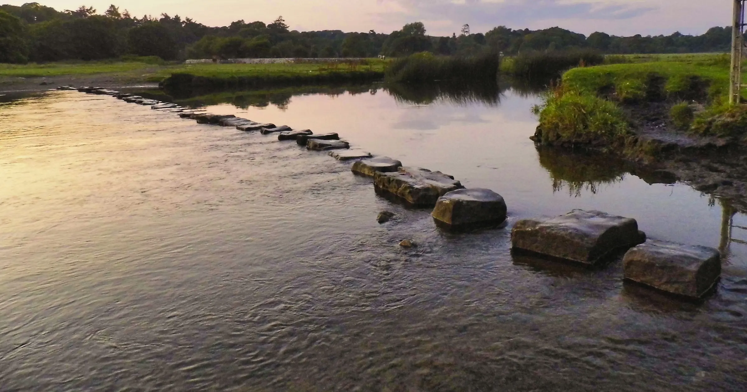 A line of flat stepping stones crossing a calm river toward a grassy bank at dusk.