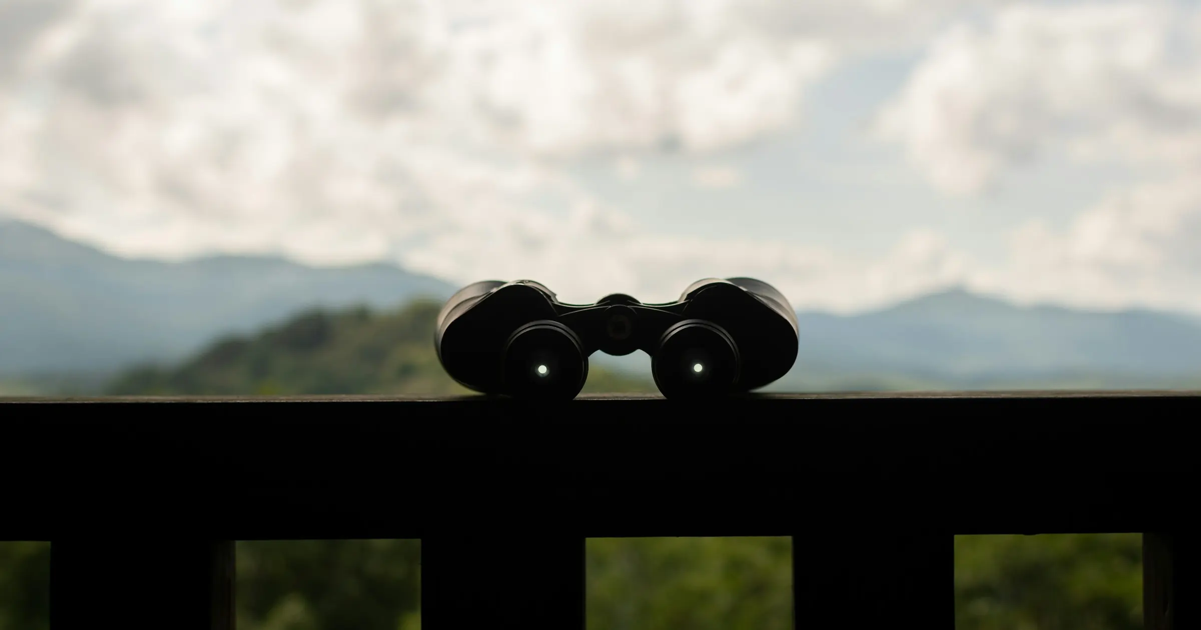 Binoculars resting on a dark wooden railing, silhouetted against hazy mountains and a cloudy sky